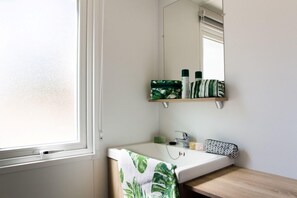 Et badeværelse med en hvid vask, et spejl og et vindue. Toiletartikler er monteret på væggen og et håndklæde ligger på vasken.