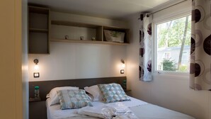 Ein ordentlich bezogenes Bett mit Kissen und einem gefalteten Handtuch, ein Nachttisch mit einer Wasserflasche und ein Fenster mit Vorhängen.