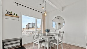 A dining area with a round table, white chairs, a bench, a hanging light fixture, a mirror, and a window with a view of buildings. - Bright, new suite 10 min from Calgary (Okotoks)