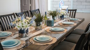 Uma mesa de jantar posta com pratos azuis, copos transparentes e um arranjo de flores e folhagens.