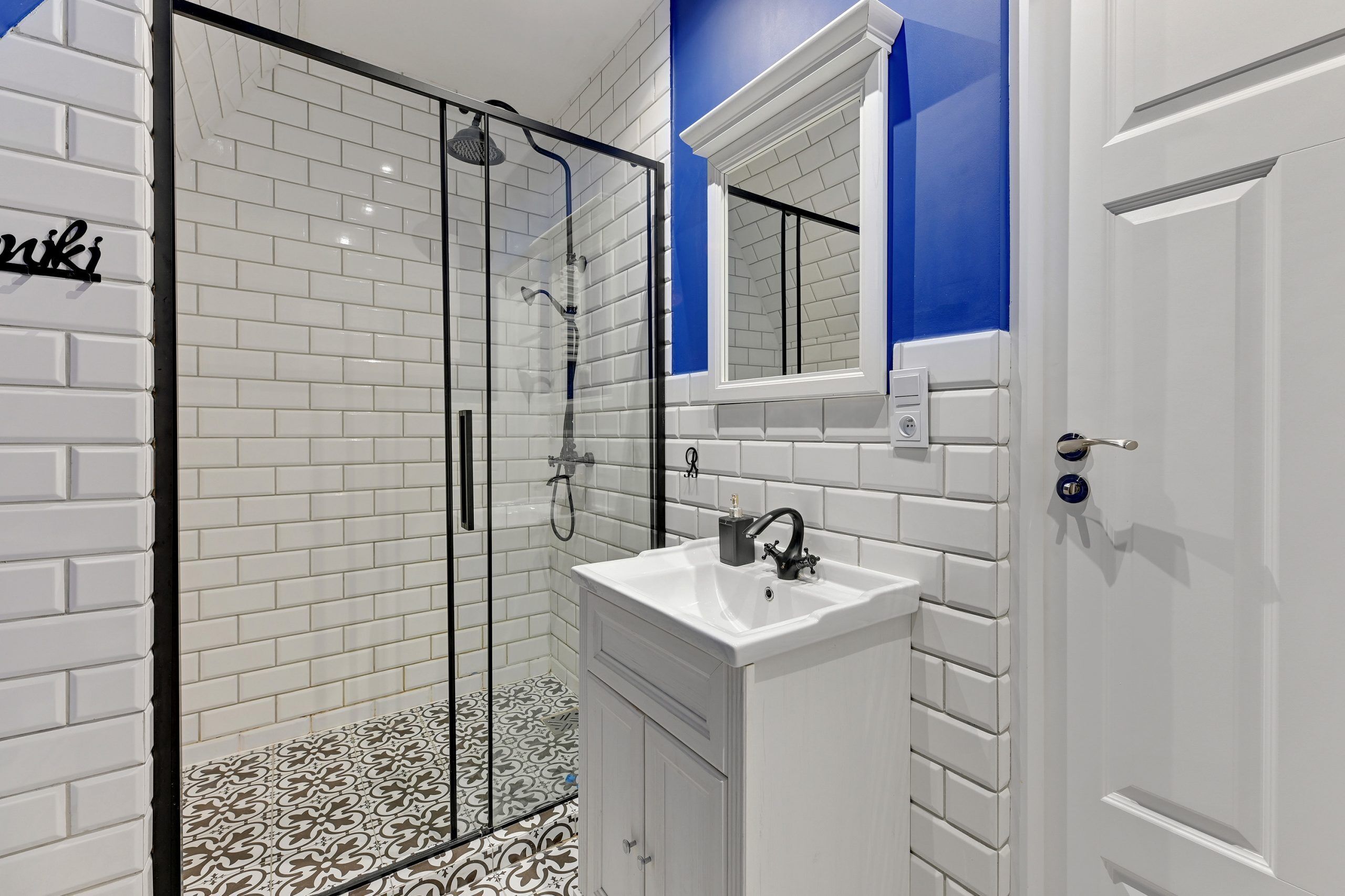 A bathroom with white tiled walls, a glass shower enclosure, a white sink, and a door with a handle.