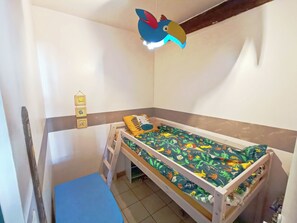 Een kleine eenpersoonskamer met een houten frame, een blauwe matras en een bedsprei met een kleurrijk bladmotief. Er is een klein ladder om een onderbed of opbergruimte te bereiken, en een wandplank met enkele spullen.