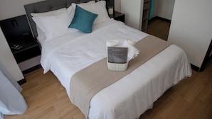 A neatly made bed with white linens and a teal pillow, a bedside table with a lamp, and a basket of toiletries on the bed.