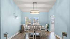 Un comedor con una mesa y sillas blancas, una lámpara colgante y dos ventanas con vistas al exterior.