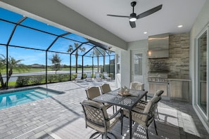 An outdoor patio with a pool, a table with chairs, a ceiling fan, and a stone fireplace. - New Home w/ Pool at The National in Ave Maria FL (Ave Maria)