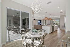 A modern kitchen with a central island, a dining area with a round table, and a living space with a sofa and a TV. - New Home w/ Pool at The National in Ave Maria FL (Ave Maria)