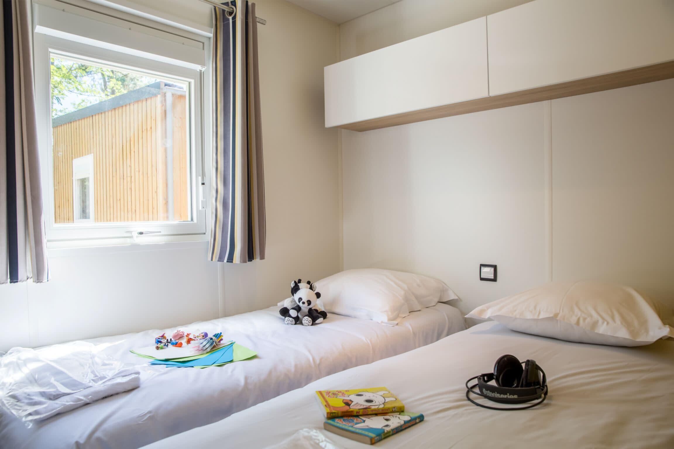 Quarto com duas camas, uma janela com cortinas, um ursinho de panda e um par de fones de ouvido em uma das camas.