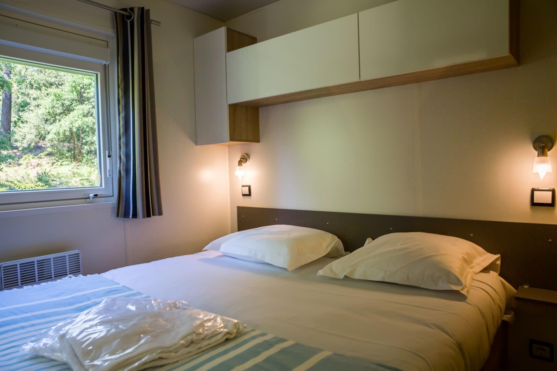 Ett hotellrum med en säng, ett fönster med gardiner, en väggmonterad lampa och ett element.