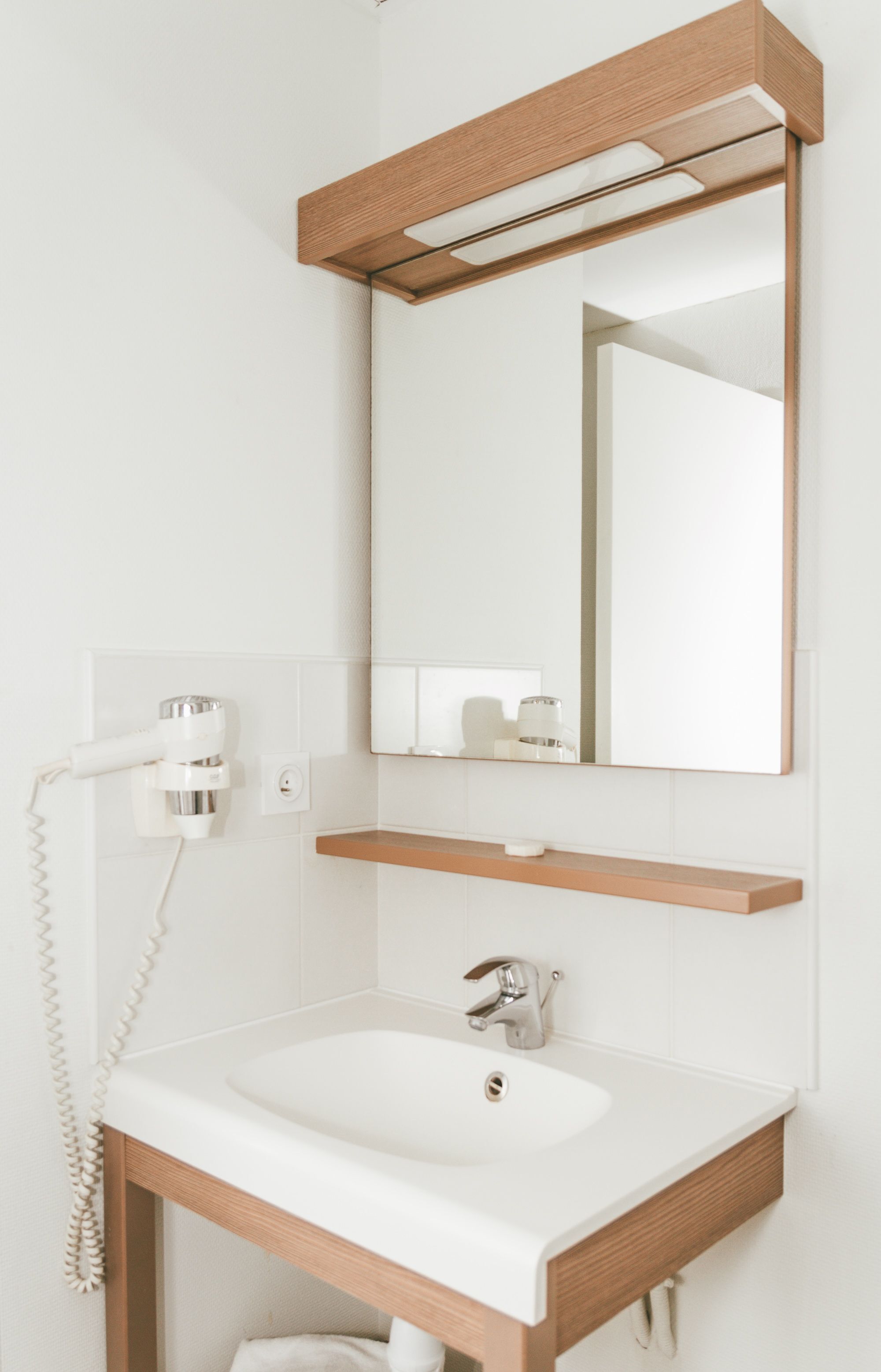 A bathroom with a white sink, wooden vanity, a mirror, and a hairdryer on the wall.