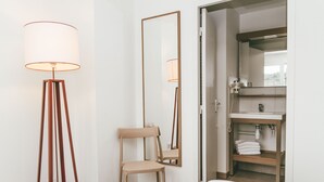Habitación de hotel con cama, lámpara de pie, silla, espejo y baño visible a través de una puerta abierta.