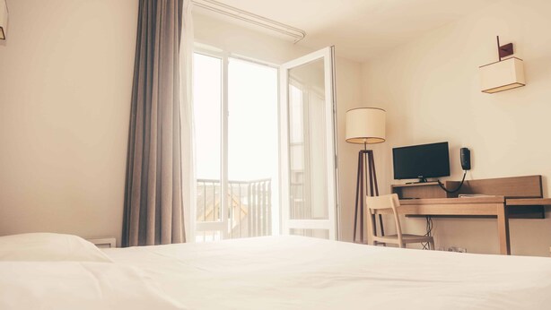 Habitación de hotel con una cama, un escritorio con computadora, una lámpara y vistas a un balcón.