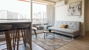 Una sala de estar moderna con un sofá gris, una mesa de centro y dos taburetes altos. Se ve un ventanal con vistas a edificios.