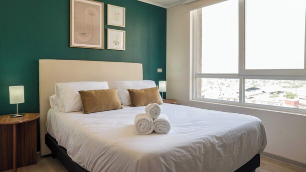 Una cama bien hecha con sábanas blancas y almohadas marrones, una lámpara de noche y una ventana con vistas a edificios.