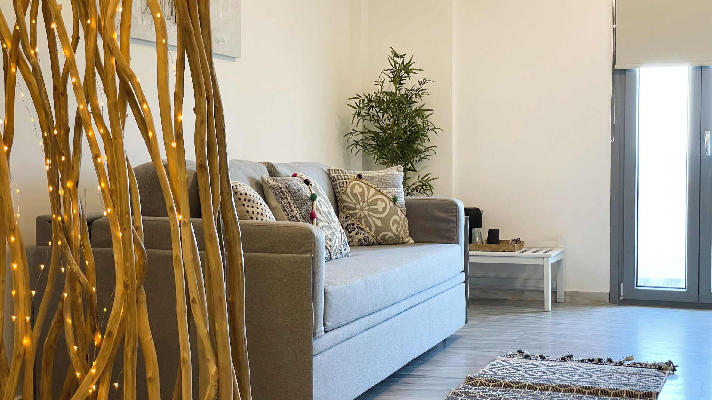 A modern living room with a grey sofa, patterned cushions, a small side table, a potted plant, and a decorative branch with lights.