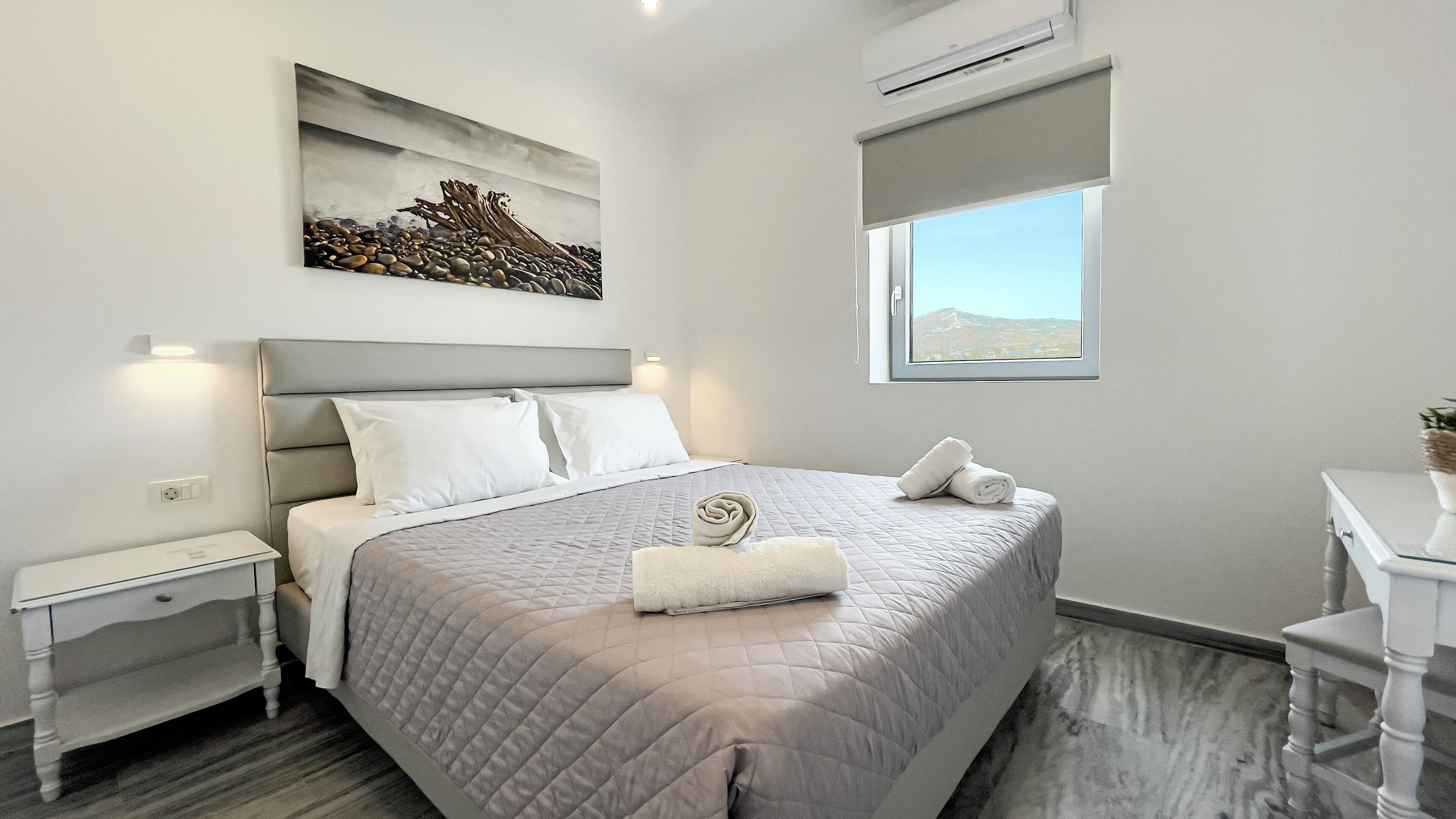 A modern bedroom with a large bed, a bedside table, a nightstand, a painting, a window, and an air conditioning unit.