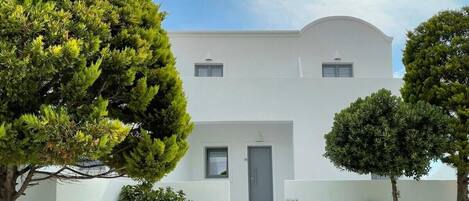A modern white building with a central entrance, surrounded by trees and a small garden.