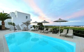 En swimmingpool med liggestole og parasoller, en trappe op til en hvid bygning og udsigt til havet.