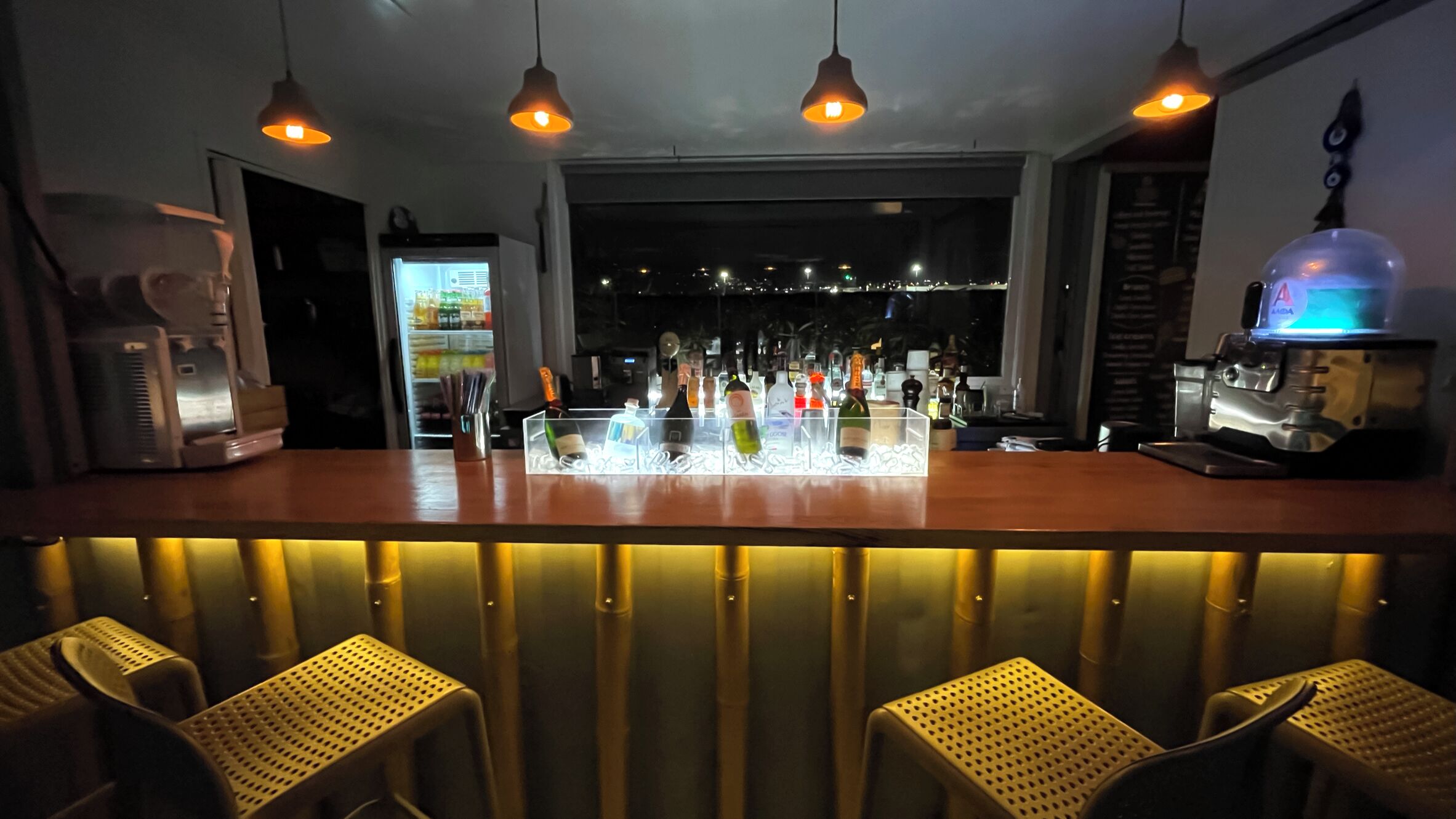 A bar area with a wooden counter, hanging pendant lights, and a view of the city at night.