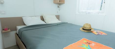 A neatly made bed with a grey bedspread, a straw hat, and a colorful pineapple pillow on a wooden bed frame.