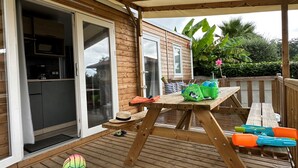 A wooden deck with a table and chairs, a sliding glass door leading to a kitchen, and a view of a house in the background. - Bela - Confort 2 TV – 5p (5) (Anglet)