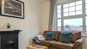 A living room with a brown leather sofa, a wooden coffee table, a fireplace, and a window with a view of buildings outside. - Digby house flat 2 (West Mersea)