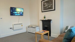 A living room with a flat-screen TV mounted on the wall, a fireplace, a couch, and a small table with cards. - Digby house flat 2 (West Mersea)