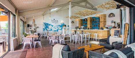 Un restaurant doté d’un bar, de chaises blanches, de meubles en osier et d’une fresque murale.