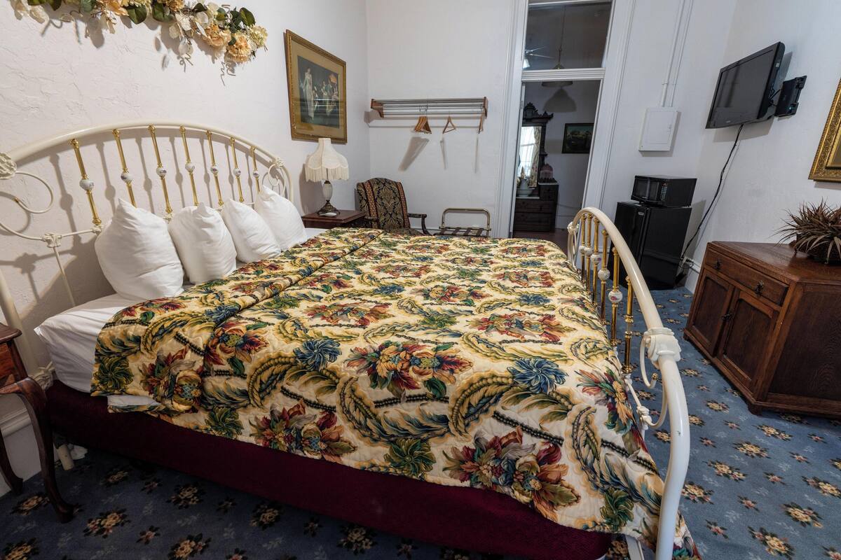 A bedroom with a floral bedspread, a white headboard, a wooden nightstand, a bedside table with a lamp, a chair, a dresser, and a television mounted on the wall.