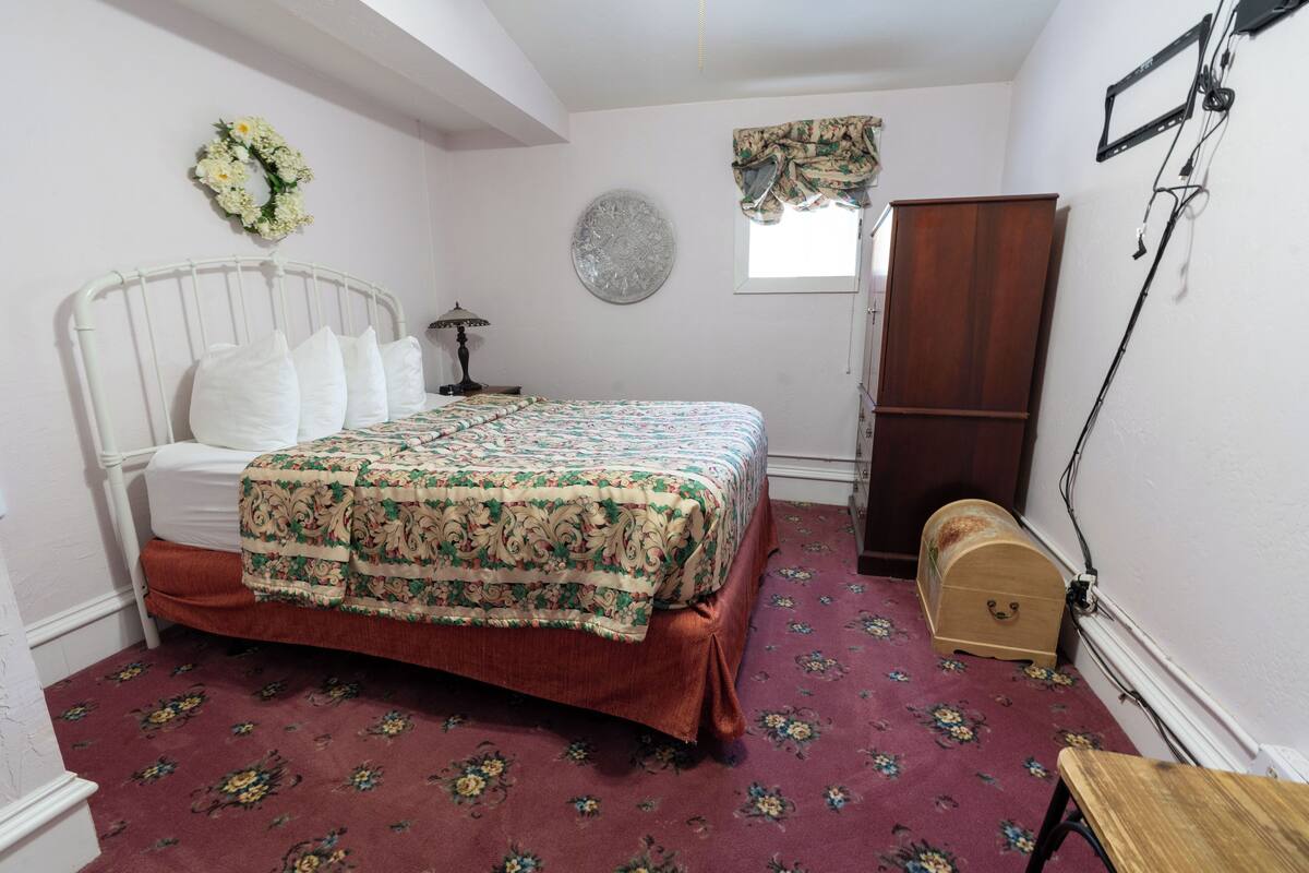 A bedroom with a bed, a wooden cabinet, a small table, a wall clock, and a window with curtains.
