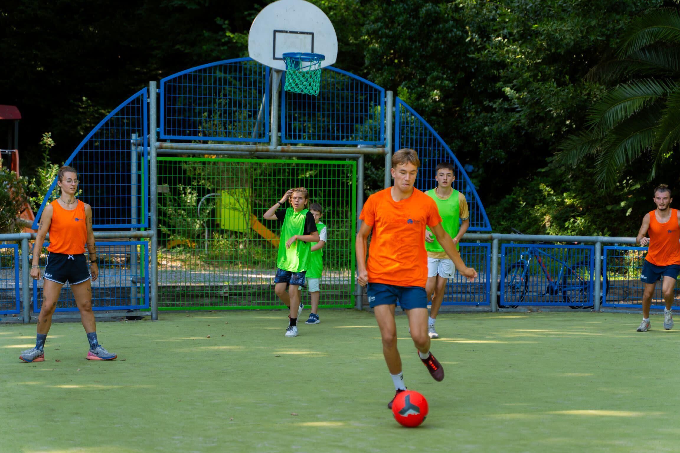 Eine Gruppe von Menschen spielt Fußball auf einem Außenplatz, im Hintergrund ist ein Basketballkorb zu sehen.