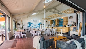 Een restaurant met een bar, witte stoelen, rieten meubels en een muurschildering.