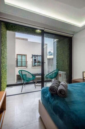 A modern bedroom with a large bed, a sliding glass door leading to a balcony, and two green chairs. - Rooftop Pool Condo Steps from 5th Ave & Beach (Playa del Carmen)
