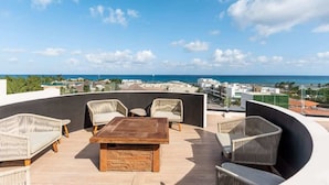 A rooftop terrace with wicker furniture, a wooden table, and a view of the ocean and cityscape. - The Boat King Studio 200m to Beach w Rooftop Pool (Playa del Carmen)