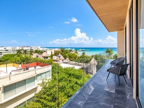 A balcony with a view of a beach and ocean, a seating area with chairs, and a building with a glass facade. - 1BR in Singular Joy, best location in town (Playa del Carmen)