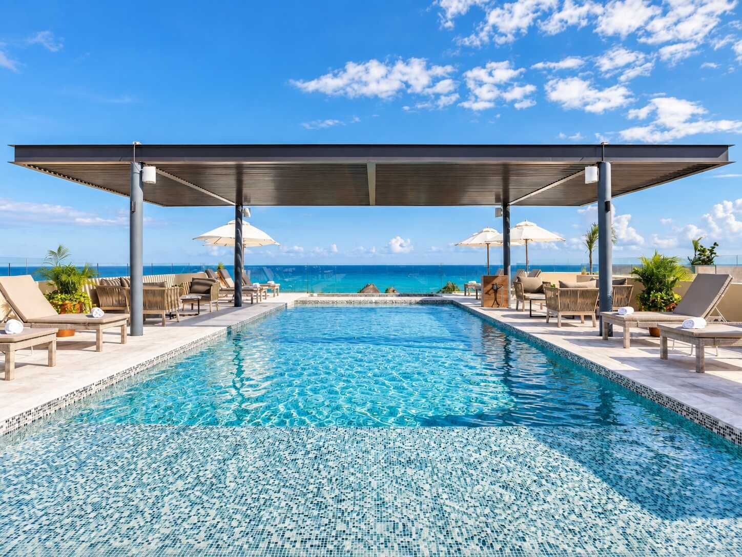 Ein Pool mit Meerblick, Liegestühle, Sonnenschirme und ein überdachter Bereich.