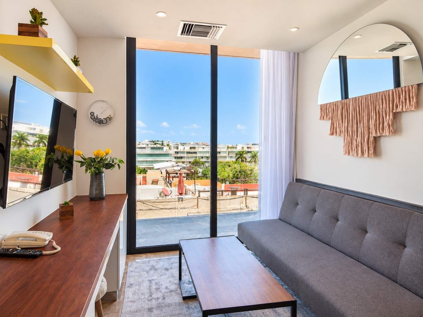 Quarto de hotel moderno com sofá, escrivaninha com telefone, espelho grande e vista para a cidade pela sacada.