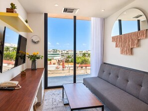 A modern hotel room with a sofa, a desk with a phone, a large mirror, and a view of the cityscape through a balcony. - 1BR in Singular Joy, best location in town (Playa del Carmen)