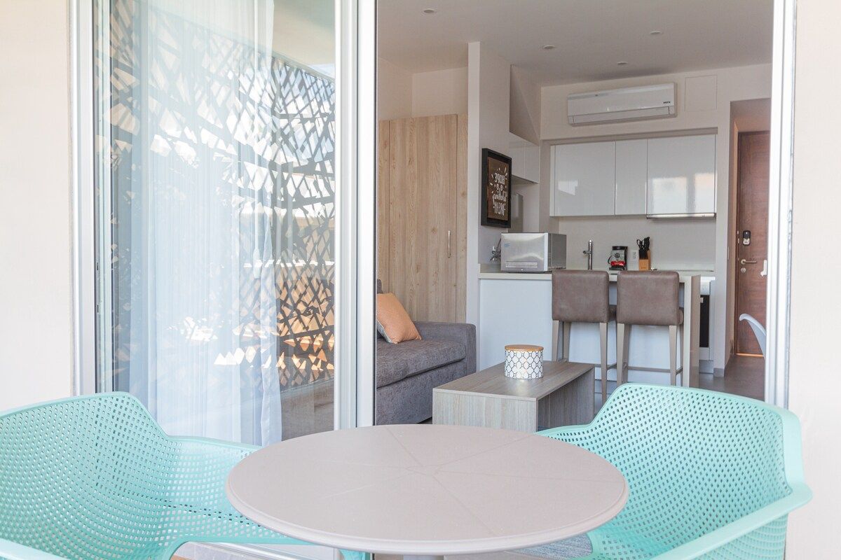 Una sala de estar moderna con una mesa redonda y dos sillas verdes, un sofá gris y una cocina con armarios blancos y un microondas.