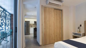 Et moderne hotelværelse med en træskab, en seng med mørkt sengegavl, et lille natbord med lampe, og udsigt til et tekøkken.