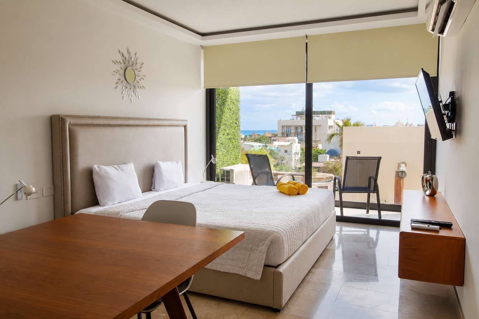 Ett hotellrum med en säng, ett skrivbord, en stol, en TV och en balkong med utsikt över byggnader och havet.