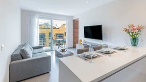 Een moderne woonkamer met een grijze bank, een eethoek met een witte tafel gedekt voor twee personen, en een aan de muur bevestigde televisie.