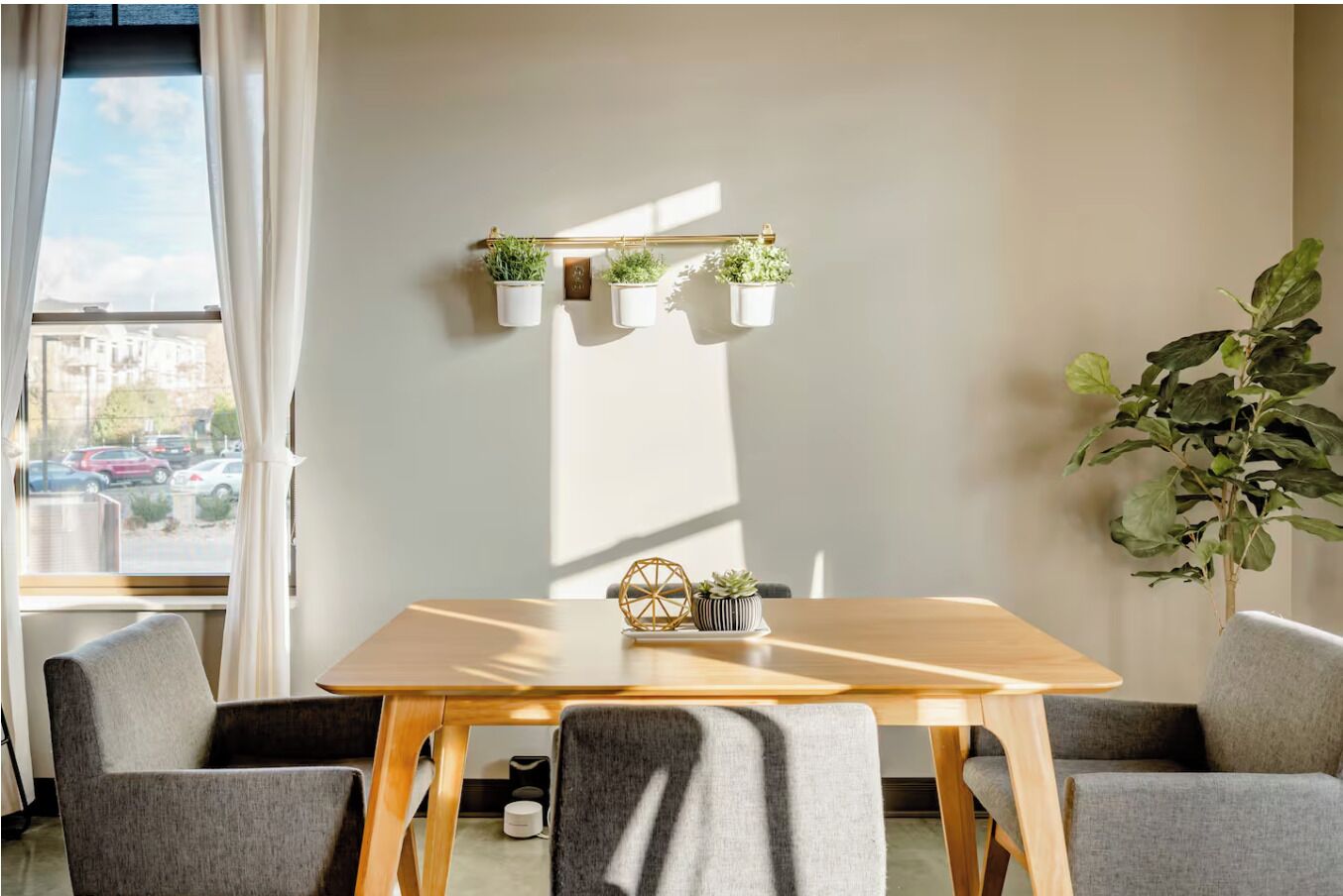 Une pièce moderne avec une table en bois, quatre chaises grises, une fenêtre avec des rideaux blancs et une étagère murale où sont disposées cinq plantes.