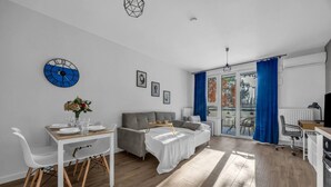 Een moderne woonkamer met een eethoek, een grijze bank, een wit bed en een groot raam met blauwe gordijnen.