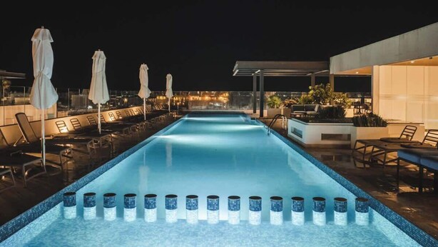 Ein Pool bei Nacht mit Liegestühlen und Sonnenschirmen, umgeben von moderner Architektur.