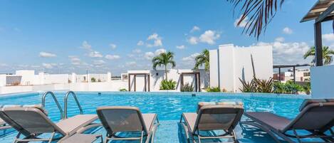 Un espace piscine avec des chaises longues, un ciel d’un bleu éclatant et des palmiers.
