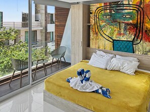 Een hotelkamer met een groot raam, een bed met een geel dekbed, een schilderij aan de muur, en een balkon met stoelen en een tafel.