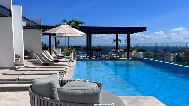 Zona de piscina con sillas de descanso y una zona de estar cubierta, con vista a un cielo azul despejado y un paisaje distante.