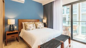 Habitación de hotel con cama, lámparas de noche, ventilador de techo, ventana con cortinas y balcón con sillas.