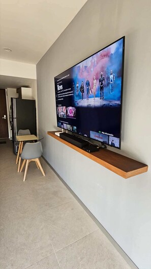 A modern living room with a large flat-screen TV mounted on a wall, a wooden shelf below, and a small table with two chairs. - Modern Studio w Pools & Gym Near 5th Ave (Playa del Carmen)