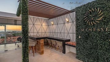 Un bar en la azotea con un diseño geométrico en la pared, una pérgola de madera y vistas de la ciudad.